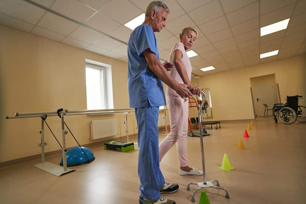 Health care worker helps female patient with adjustable walking cane restore mobility skills during exercising in rehab center